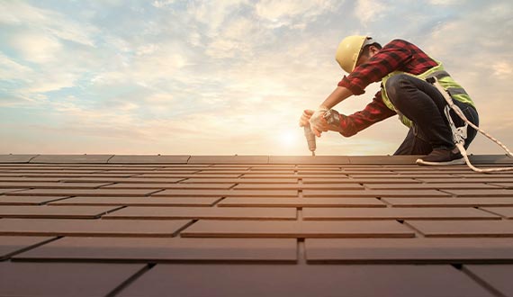 roofer working in special protective work wear gloves using air or pneumatic nail gun installing roofing tiles on residential building